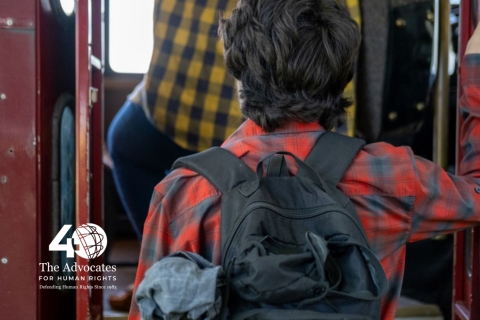 [ai] A young person wearing a red plaid shirt and carrying a black backpack is depicted from behind as they board a bus. Another passenger wearing a yellow and blue checkered shirt is seen in the background.